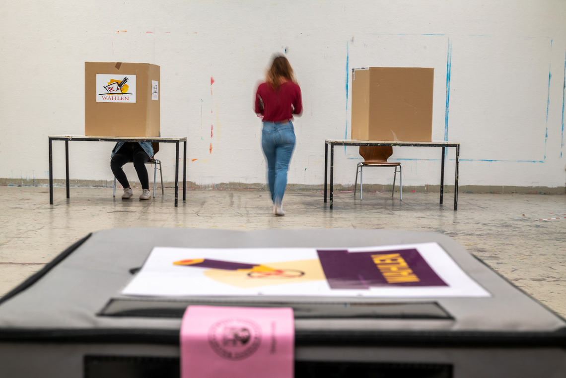 Junge Frau geht zwischen zwei Wahlkabinen, im Vordergrund Wahlunterlagen