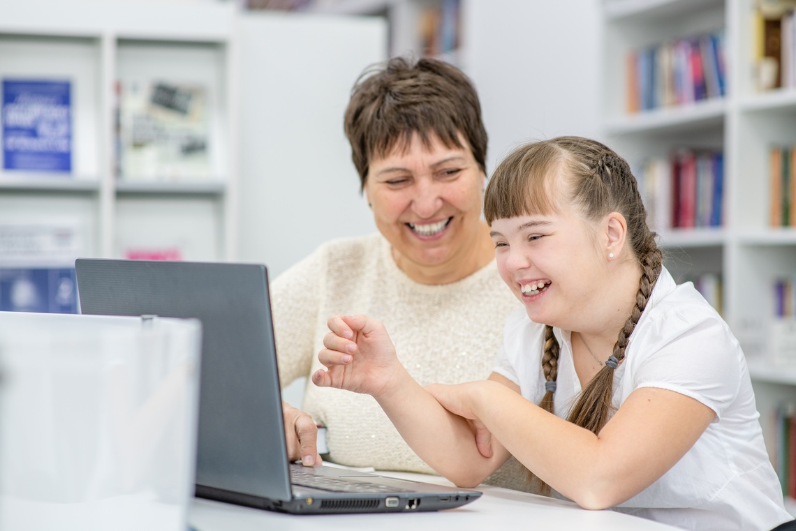 Frau und Kind mit Behinderung lachen gemeinsam vor einem Laptop.