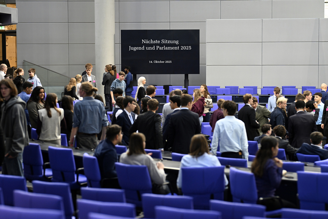 Jugendliche im Plenarsaal mit blauen Sesseln und einem Bildschirm mit "Jugend und Parlament"