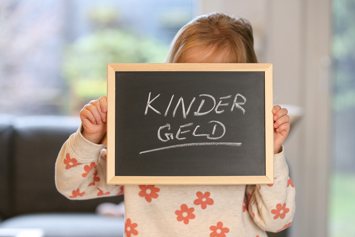 Kind hält kleine Schiefertafel in den Händen, das Gesicht wird davon bedeckt, weiße Kreide zeigt das Wort Kindergeld