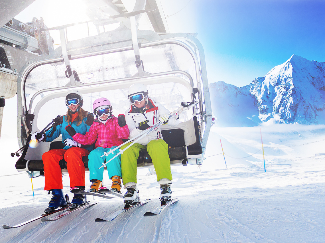 Drei Personen sitzen im Skilift. Im Hintergrund verschneite Berge.