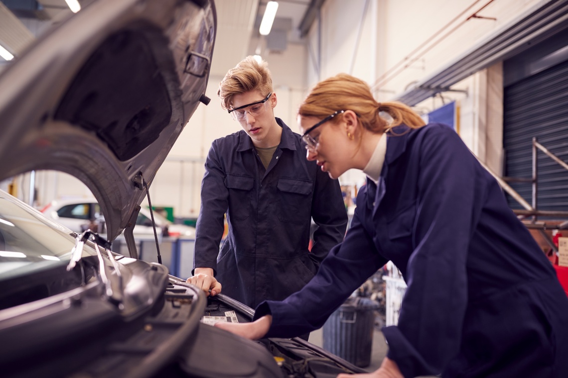 Junge Frau und junger Mann in Arbeitskleidung schauen in eine Automotorhaube.