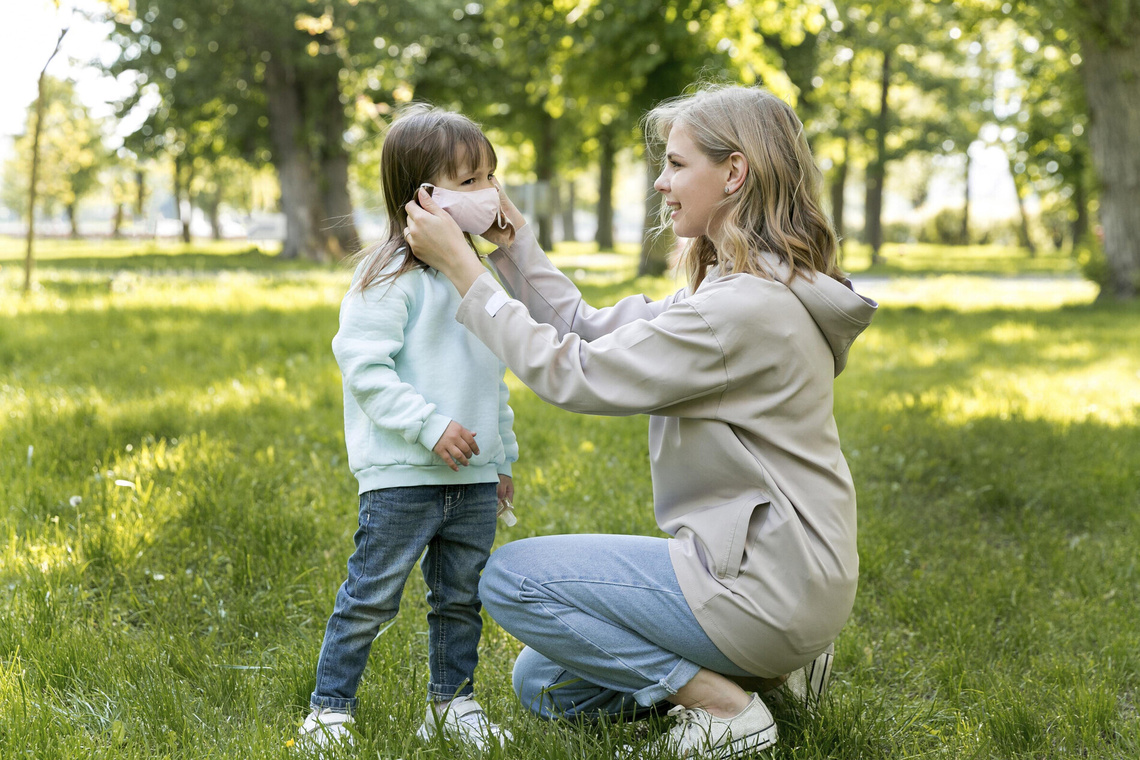 Ein Foto einer Mutter, die ihrer kleinen Tochter im sonnigen Park eine rosa medizinische Maske anlegt.