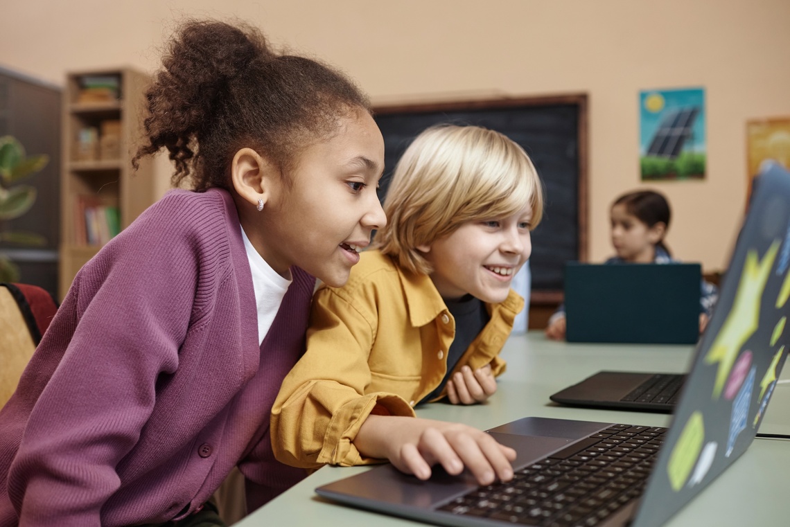 Junge und Mädchen sitzen am Tisch und schauen auf einen Laptop.