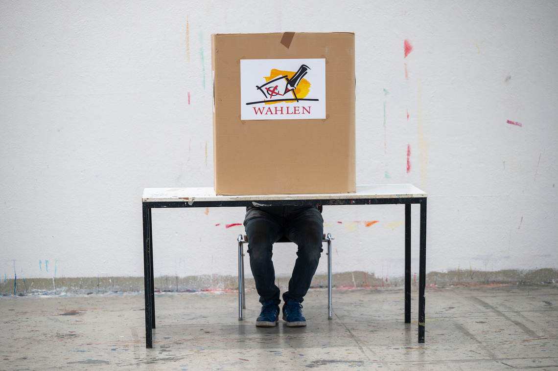 Ein Mann sitzt an einem Tisch, sein Oberkörper ist von der Wahlkabine verdeckt. Darauf ein Plakat "Wählen!"