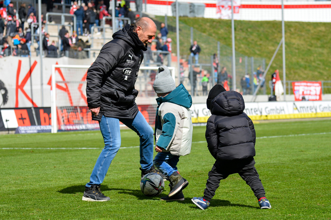 Fußballplatz mit einem Trainer und zwei kleinen Kindern in dicken Jacken spielen Fußball