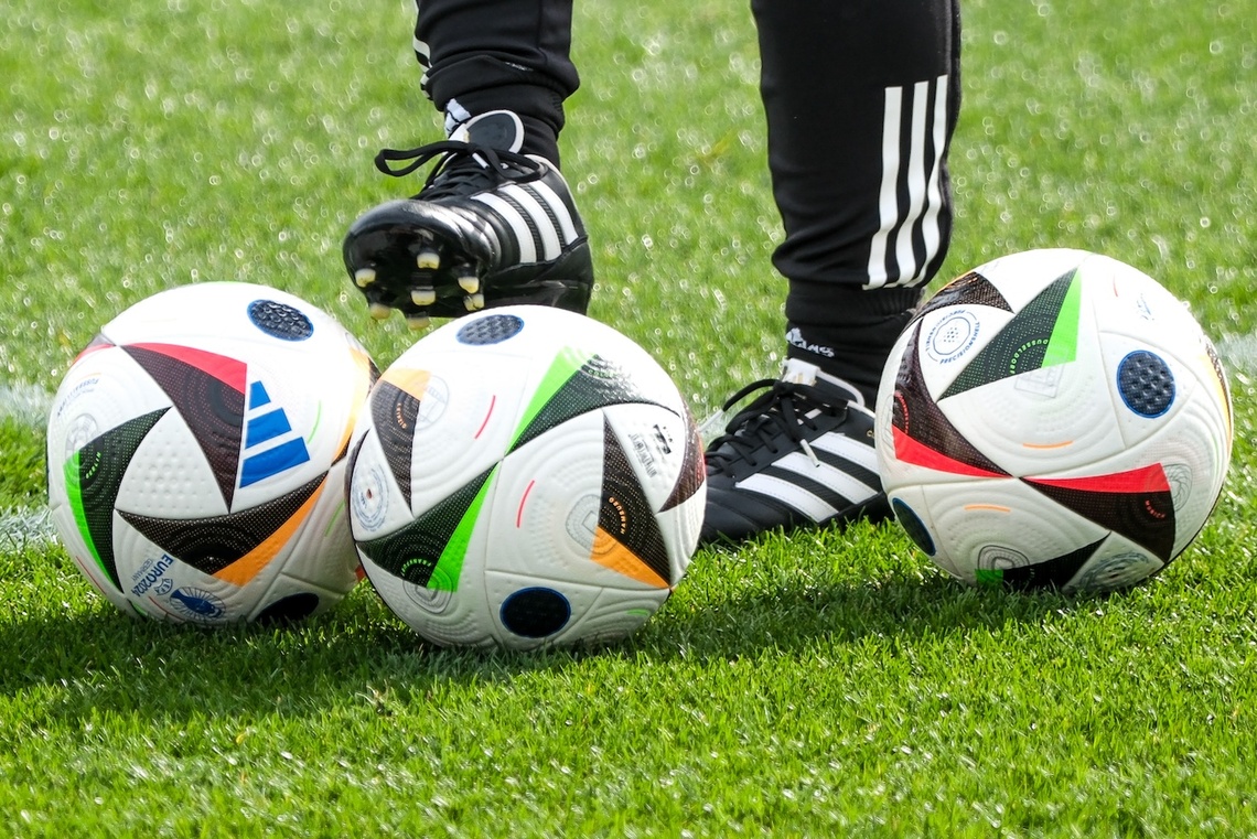 Drei Fußbälle liegen auf dem Rasen. Im Hintergrund sind Beine in Fußballschuhen.