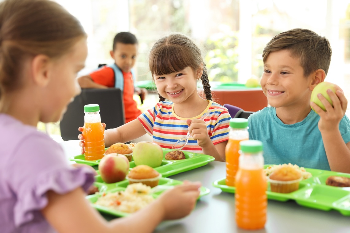 Lächelnde Kinder am Tisch beim Mittagessen. Auf dem Tisch sind Saft und Lebensmittel.