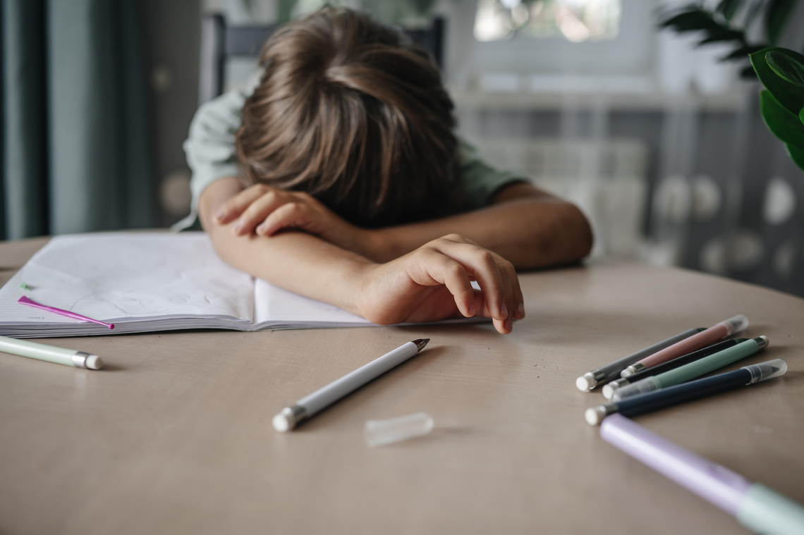 Kind liegt mit Kopf auf dem Tisch über Schulmaterial