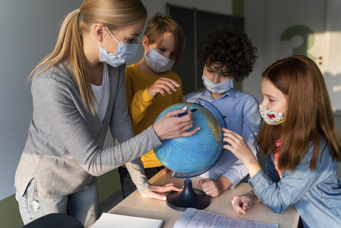 Lehrerin zeigt Kindern Orte auf einem Globus, alle tragen Mundschutz