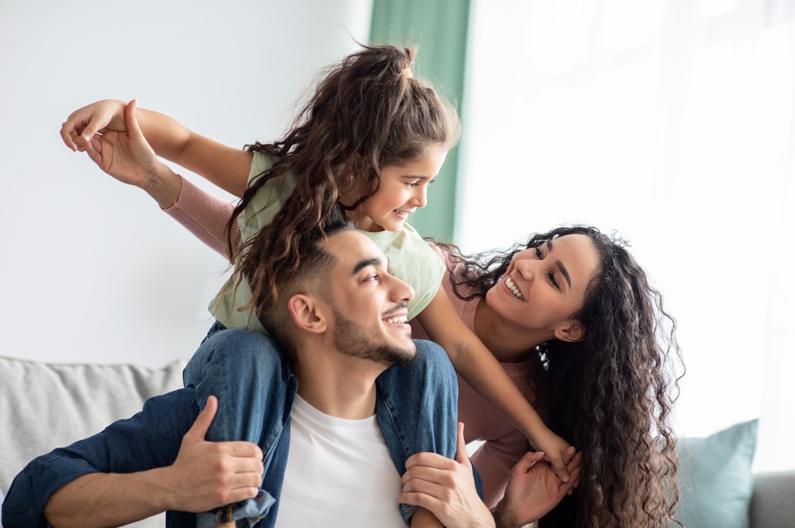 Vater mit Tochter auf den Schultern lächelt seine Frau an.