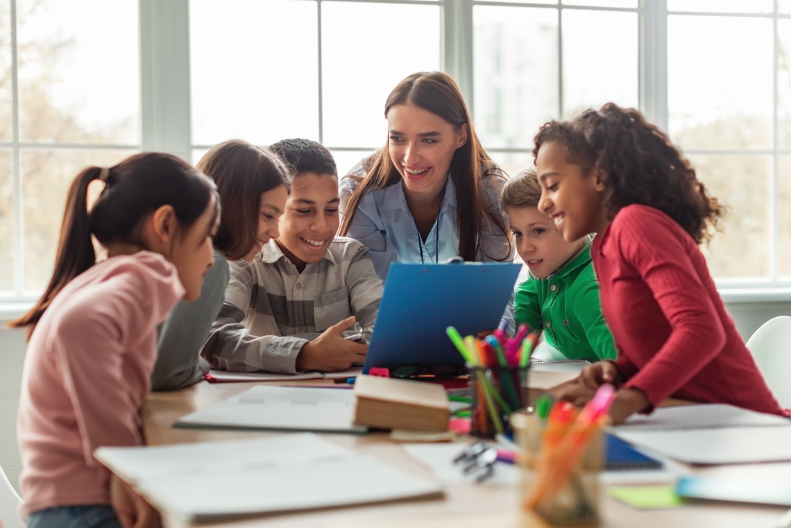 Lehrerin sitzt mit fünf Kindern im Halbkreis an einem Tisch.