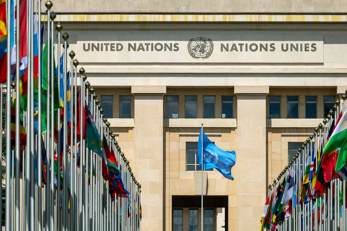 Hauptgebäude des Palais des Nations (Büro der Vereinten Nationen in Genf) am Place des Nations. Die helle, sandsteinfarbene Fassade trägt großflächig die Inschrift "UNITED NATIONS" und "NATIONS UNIES" sowie das UN-Emblem. Vor dem Gebäude stehen zwei symmetrische Reihen hoher Fahnenmasten mit zahlreichen bunten Nationalflaggen; in der Bildmitte weht auf einem separaten Mast eine blaue Flagge mit dem UN-Emblem.