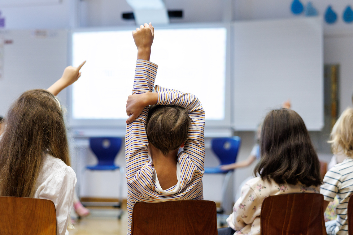 Kinder sitzen in einem Klassenraum, ein Kind zeigt auf.