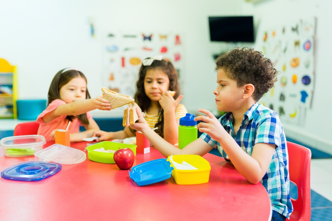 Drei Kinder sitzen am Tisch mit Brotbüchsen und tauschen Brote aus.