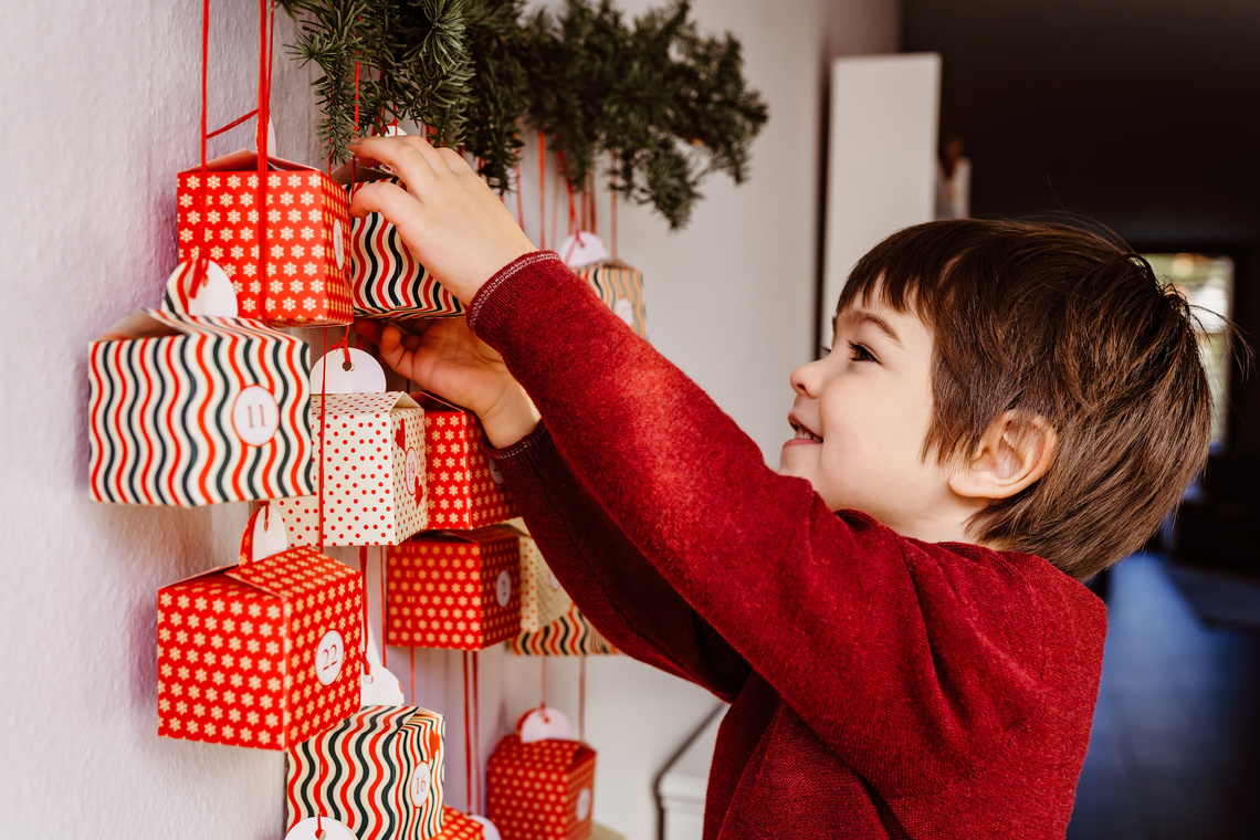 Fröhlicher Junge beginnt, einen Adventskalender zu öffnen, der an der Wand hängt.