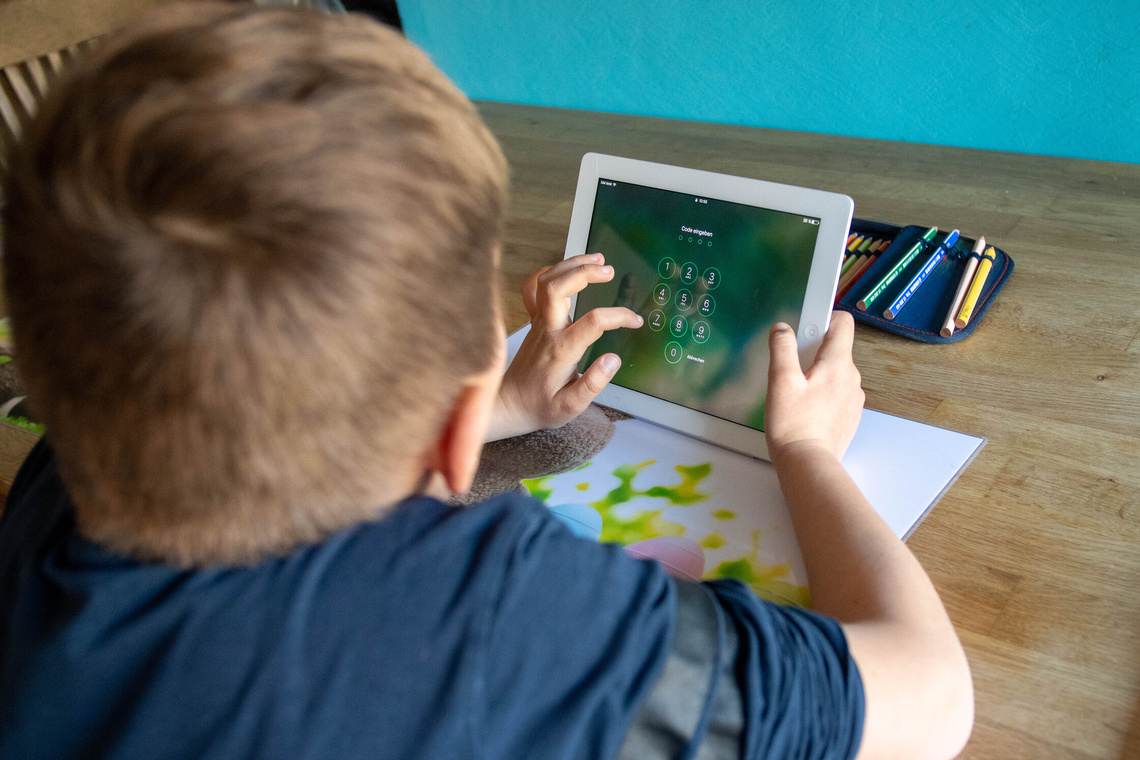 Kind von hinten fotografiert, sitzt am Schreibtisch, hat Tablet in der Hand und muss Pin-Code eingeben