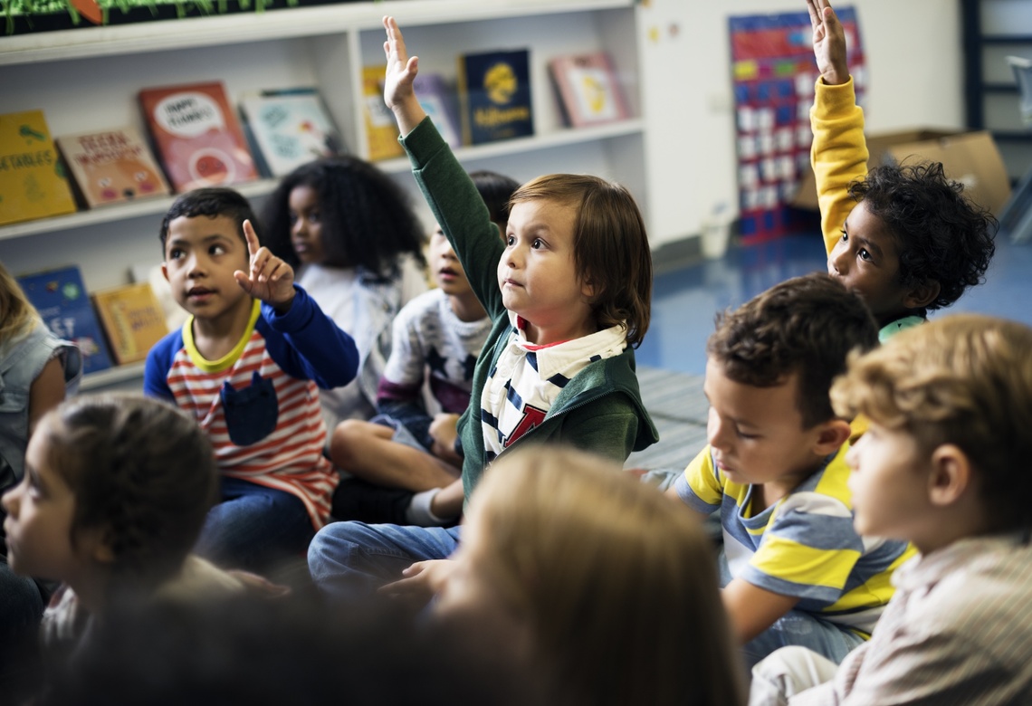 Eine Kindergartengruppe sitzt auf dem Boden. Drei Kinder melden sich.