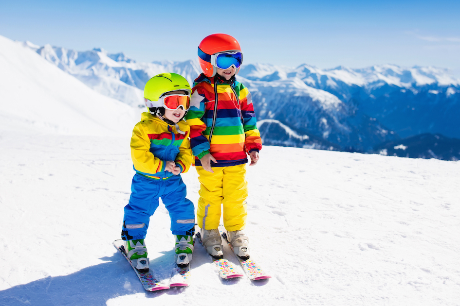 Zwei Kinder auf Skiern im Schnee. Im Hintergrund Berge.